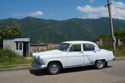 White vintage car on a country road with mountain backdrop and blue sky in summer, GAZ M-21 Volga,