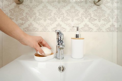 A hand reaches for a bar of soap placed on a dish near a sink. Water flows from a silver faucet