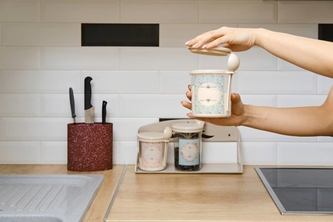 A person holds a container in one hand while opening the lid with the other. The kitchen has a