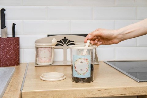 A hand is reaching into a jar with tea on a wooden kitchen countertop. Nearby, there is another jar
