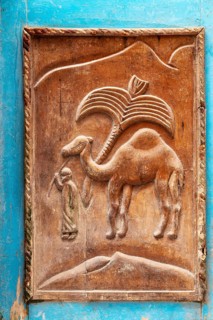 Wood carving of a camel depicting a person in traditional clothing against a hill backdrop,