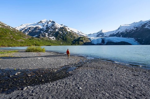 Young man on a pebble beach on a glacial lake, snowy mountains and Portage Glacier glacier on