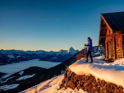 Mountaineers enjoy the sunrise in front of an alpine hut in winter, the Watzmann behind, fog in the