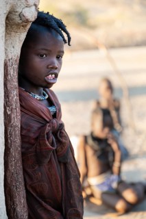 Himba girl leaning at a traditional clay hut, in the morning, traditional Himba village, Kaokoveld,