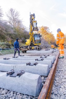 Concrete ties are laid with an excavator along a railway line, supervised by workers, track