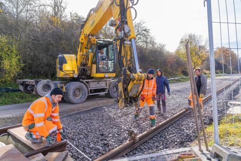 Construction workers in orange clothing work next to a yellow excavator on a railway line, track