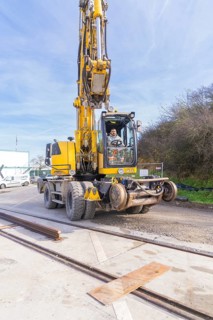 A yellow excavator on a construction site with tracks in the foreground. The sky is clear and the