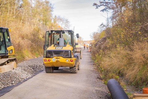 A yellow excavator is standing on a construction site, surrounded by autumn trees. Workers can be