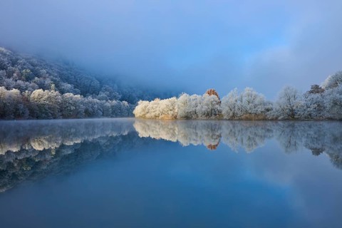 A calm river reflects frosty trees and mountains in a wintry environment, hoarfrost, winter,