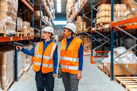 Two women wearing hard hats and safety vests are analyzing inventory by scanning product barcodes