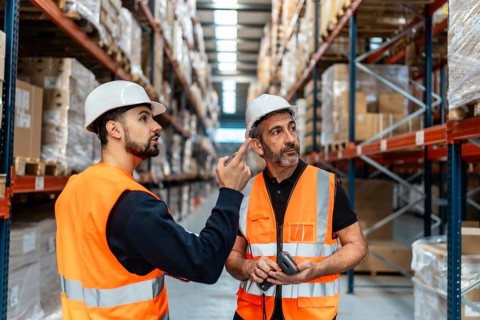 Two male workers wearing hard hats and safety vests are discussing inventory and supply chain