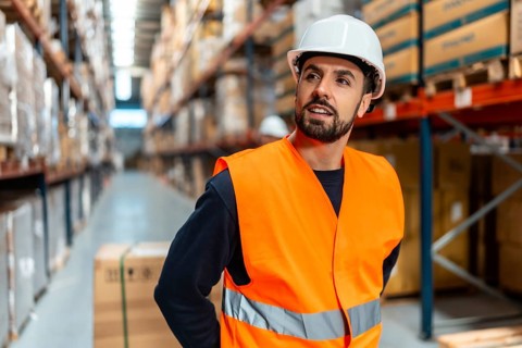Warehouse worker in hard hat and orange safety vest standing in an aisle, inspecting high shelves