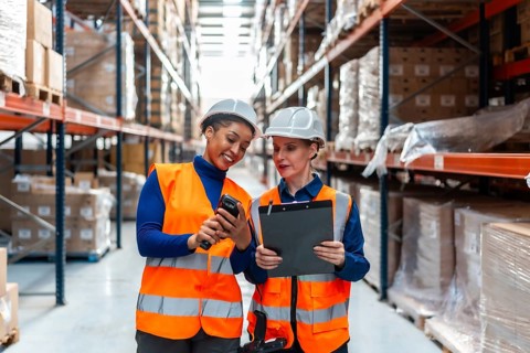 Two women warehouse workers in safety hats and vests are collaborating, scanning inventory with a