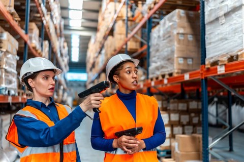 Women in hard hats and safety vests collaborate in a large warehouse, using handheld scanners to