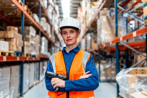 Woman wearing hard hat and safety vest standing confidently in a large logistics warehouse, holding