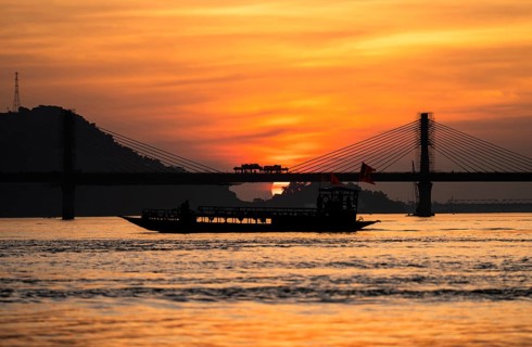 A boat sails on the river Brahmaputra during sunset, with the silhouette of a cable-stayed bridge