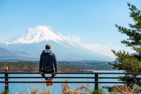 Young man sitting on railings next to a road and looking across the lake to Mt Fuji volcano, Motosu