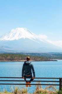 Young man sitting on railings next to a road and looking across the lake to Mt Fuji volcano, Motosu
