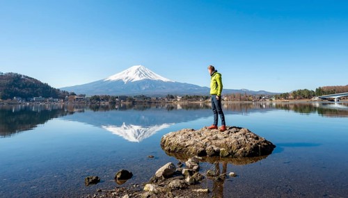 Young man standing on a rock in water, volcano Mt. Fuji is reflected in Lake Kawaguchi, Yamanashi