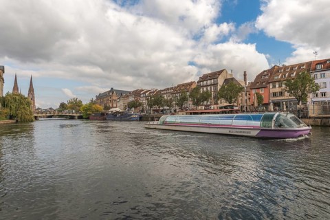 A pleasure boat glides peacefully on the Strasbourg River. Colourful buildings and trees line the