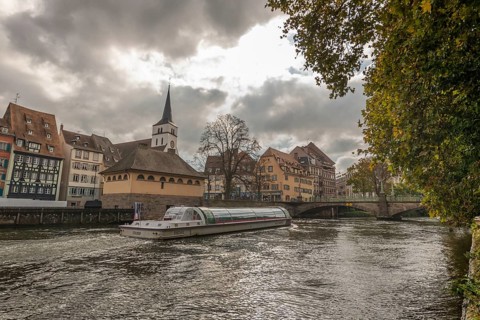 Boat sails gently on river in Strasbourg. Historic houses are located on the banks. The sky is