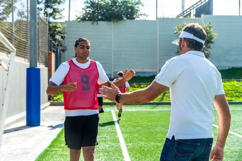 Soccer coach extends a hand to a young male player wearing a number 9 bib on a green artificial