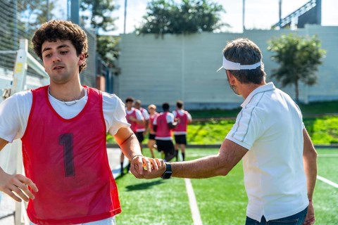 Coach instructing a soccer player on the field during training, teammates jogging in the background