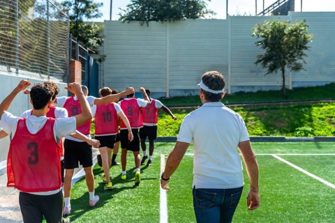 Youth soccer coach leading a group of players in red training vests across artificial grass,