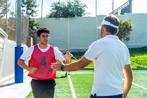 Soccer coach high fiving young male player in red bib on green artificial turf, offering