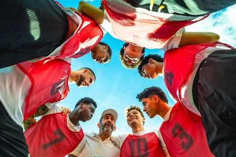 Diverse male soccer team and coach in a pregame huddle on a sunny day, sharing motivation and