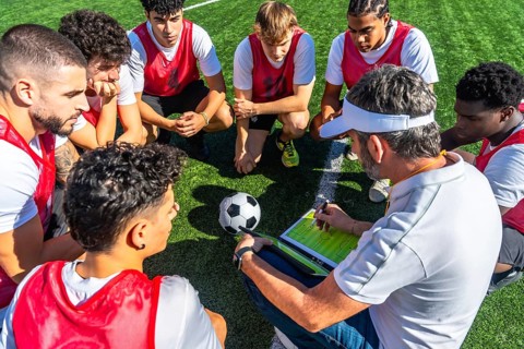 Soccer coach huddling with his youth team, drawing plays on a tactical board, and discussing