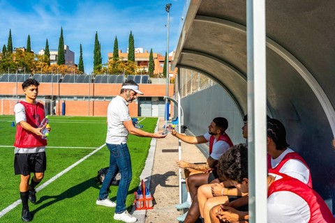 Soccer coach distributing water bottles to young male players sitting in the dugout during a