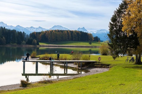 People walk along the jetty with views of Lake Forggensee and surrounding mountains, autumn trees