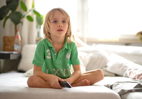 Child, boy, 7 years old, blonde, sitting on sofa, with remote control in hand, watching TV,