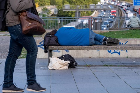 Man sleeping on a bench, travelling bag serves as pillow, in the city centre, passers-by walk by