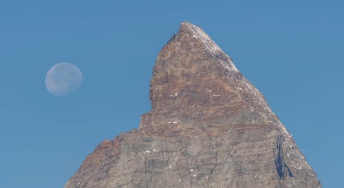 The Matterhorn mountain peak rises steeply against a bright blue sky. The full moon glistens gently
