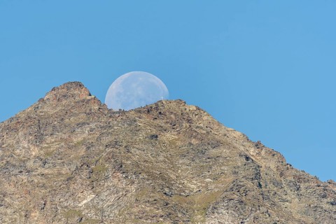 Majestic mountain peak stands tall against vibrant blue sky as large moon rises just behind it. The