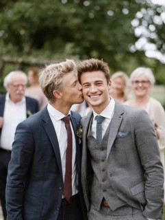 A male couple in a suit, one kissing the other on the cheek, surrounded by guests outdoors,