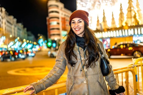 Young woman wearing winter clothes smiling in a city street decorated with christmas lights at