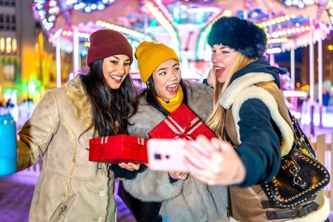 Three happy women holding christmas gifts and taking a selfie at the christmas market with a