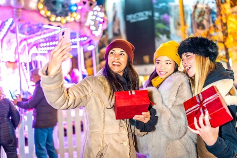 Three happy women holding christmas gifts are taking a selfie in front of a carousel in a christmas