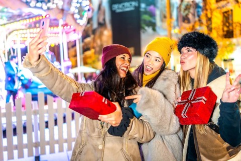 Three happy female friends are taking a selfie holding christmas gifts at a christmas market,