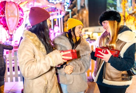 Three cheerful female friends exchanging christmas gifts at a night market with illuminated