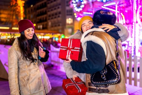 Three happy female friends exchanging christmas gifts and hugging at a christmas market at night