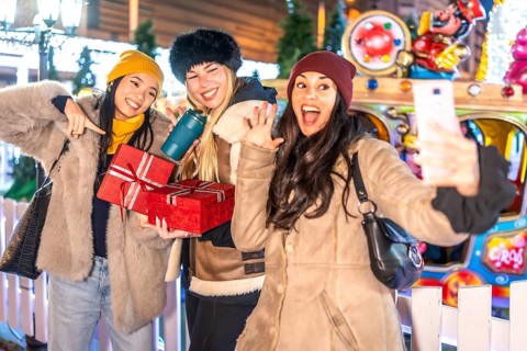 Three happy women taking a selfie at christmas market, holding presents and enjoying winter