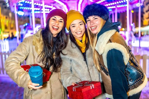 Three happy female friends enjoying christmas market at night, holding gifts and hot beverages,