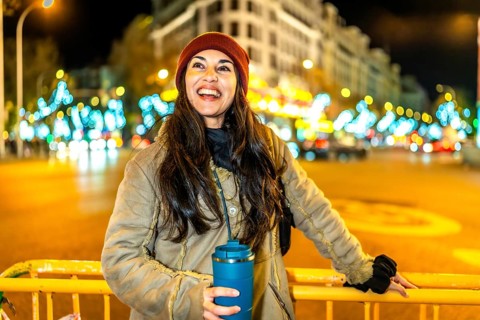 Young woman holding reusable mug smiling near christmas lights decorations in a city at night
