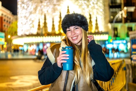 Smiling woman with winter clothes and hot beverage enjoying christmas lights and attractions in a
