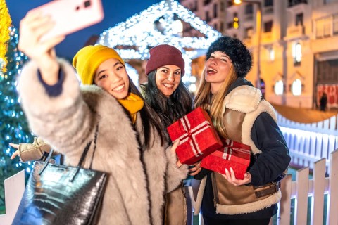 Three cheerful women holding christmas gifts taking a selfie with smartphone at christmas market in