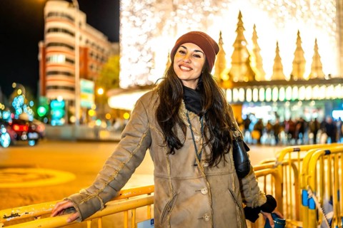 Smiling young woman leaning on a fence in a city street enjoying christmas lights and decorations